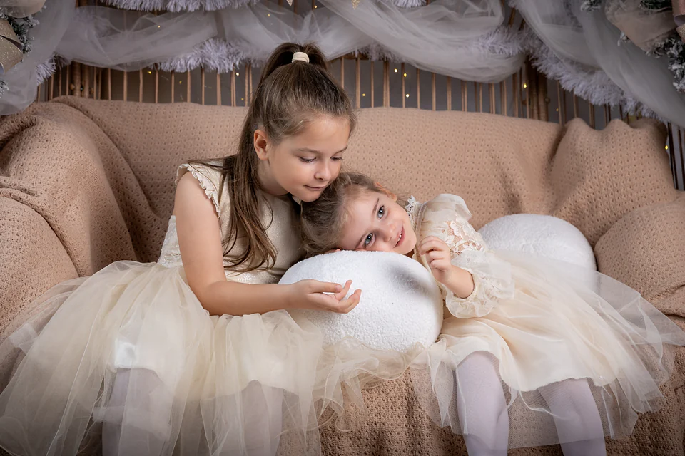 Două surori în rochițe ivoire, îmbrățișate pe canapeaua de răchită din decorul de Crăciun alb și auriu — ședință foto de Crăciun în studio, Oradea