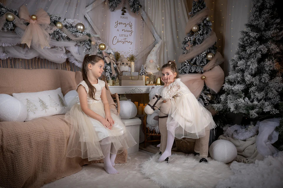 Fetiță pe un călușel de lemn și sora ei lângă bradul nins, în decorul alb și auriu — studio foto de Crăciun Oradea, Bihor