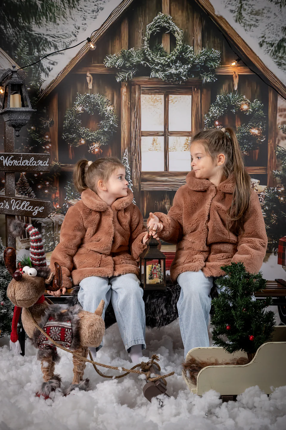 Surori care se privesc ținând un felinar aprins, în decorul de iarnă cu căsuță de lemn — fotografie de Crăciun pentru copii, Oradea
